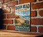 A vintage-style metal sign for "Basin Head" in Prince Edward Island, featuring a beach scene with a sign for "Singing Sands" and "Nature's Music A Maritime Treasure".