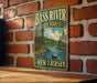 A vintage-style metal sign for Bass River State Forest in New Jersey, featuring a scenic illustration of a river surrounded by trees and mountains.