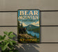 A vintage-style metal sign for Bear Mountain State Forest in New Hampshire, featuring a black bear standing on a rocky outcropping overlooking a lake surrounded by pine trees.