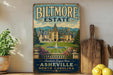 A vintage-style metal sign for Biltmore Estate in Asheville, North Carolina, featuring an illustration of the estate's gardens and fountain.