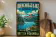 A vintage-style sign for Birkenhead Lake, a scenic park in British Columbia, Canada. The sign features a mountain landscape with pine trees and a lake, and is displayed on a wooden shelf next to a wooden cutting board and some lemons.