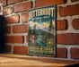 A vintage-style metal sign for Bitterroot National Forest, featuring a mountain landscape with pine trees and a river, mounted on a brick wall.