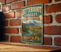 A vintage-style metal sign for the Bitterroot Valley in Montana, featuring a scenic landscape with mountains, a river, and a small town.