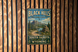 A vintage-style metal sign for the Black Hills National Forest, featuring a scenic mountain landscape with a winding road and a lake.