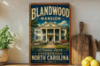 A vintage-style sign for the Blandwood Mansion in Greensboro, North Carolina, featuring a large illustration of the mansion surrounded by trees and a well-manicured lawn.