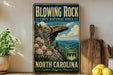 A vintage-style metal sign for Blowing Rock State Natural Area in North Carolina, featuring a scenic mountain landscape with a cliff and flowers.