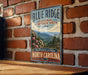 A vintage-style metal sign for the Blue Ridge National Heritage Area in North Carolina, featuring a scenic mountain landscape with pine trees and pink flowers.