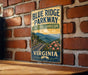 A vintage-style metal sign for the Blue Ridge Parkway in Virginia, featuring a scenic mountain landscape with a winding road and colorful flowers.