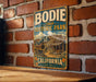 A vintage-style metal sign for Bodie State Historic Park in California, featuring an illustration of a small building and a horse-drawn carriage.