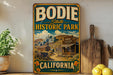 A vintage-style metal sign for Bodie State Historic Park in California, featuring a scenic illustration of a small town with a wooden building and a sign that reads "Bode California".