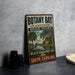 A vintage-style metal sign for Botany Bay Plantation Heritage Preserve, featuring a scenic illustration of a bird and palm trees, is displayed on a gray countertop.
