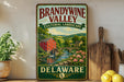 A vintage-style sign for Brandywine Valley, featuring a scenic landscape with a red brick building, a bridge, and a river with pink flowers.