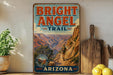 A vintage-style metal sign for the Bright Angel Trail in Grand Canyon National Park, Arizona, featuring a scenic illustration of a hiker on a trail surrounded by the Grand Canyon landscape.