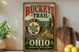 A vintage-style metal sign for Buckeye Trail, featuring a scenic illustration of a forest and a trail, is displayed on a shelf next to a wooden cutting board and some lemons.