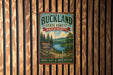 A vintage metal sign for Buckland State Forest in Massachusetts, featuring a scenic landscape with pine trees, a lake, and mountains.