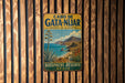 A vintage metal sign with a scenic illustration of a coastal landscape, featuring a palm tree, mountains, and a body of water, mounted on a wooden wall.