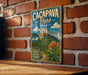 A vintage-style metal sign for Cacapa Park in Brazil, featuring a mountain landscape with orange flowers, is displayed on a brick wall.