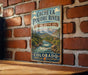 A vintage-style metal sign for the Cache La Poudre River National Heritage Area in Colorado, featuring a scenic mountain landscape with a flowing river.