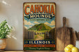 a vintage-style sign for Cahokia Mounds, a historic site in Illinois, with a mountain and a fence in the background.