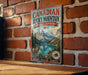 A vintage-style metal sign for Canadian Rocky Mountain Parks Canada hangs on a brick wall, featuring a scenic mountain landscape with a lake and a bighorn sheep.