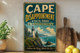 a vintage-style sign for Cape Disappointment State Park in Washington, Washington, with a lighthouse and ocean scene depicted on it. The sign is displayed on a shelf next to a wooden cutting board and a potted plant.