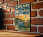 A vintage-style metal sign for Cape May Coastal Wetlands, featuring an illustration of a bird and a lighthouse, mounted on a brick wall.