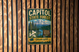 A vintage metal sign for Capitol State Forest in Washington, featuring a mountain landscape with white flowers and a river.