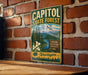 A vintage-style metal sign for Capitol State Forest in Washington, featuring a mountain landscape and a sign that says "Explore Discover Protect Washington".