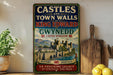 A vintage-style sign for the Castle and Town Walls of Gwynedd, United Kingdom, featuring a castle and a river.