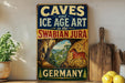 a vintage-style sign advertising "CAVES and ICE AGE ART in the SWABIAN JURA" in the Swabian Jura, Germany. The sign is displayed on a wooden shelf, with a potted plant and a wooden cutting board visible in the background.