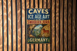 A vintage metal sign with the text "CAVES and ICE AGE ART in the SWABIAN JURA" and an illustration of a cave with a horse, hanging on a wooden wall.