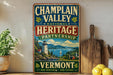 A vintage-style metal sign for the Champlain Valley National Heritage Partnership, featuring a scenic landscape with a lighthouse and mountains, is displayed on a shelf next to a wooden cutting board and some lemons.