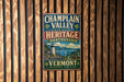 A vintage metal sign for the Champlain Valley Heritage Partnership, featuring a scenic landscape with a lighthouse and mountains, mounted on a wooden wall.
