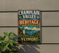 A vintage-style metal sign for the Vermont Heritage Partnership, featuring a lighthouse and a scenic landscape, mounted on a wall.