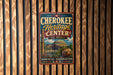 A vintage-style metal sign for the Cherokee Heritage Center in Oklahoma, featuring a colorful illustration of the center's architecture and surrounded by decorative elements.