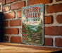 A vintage-style metal sign for Cherry Valley State Forest in New York, featuring a scenic illustration of cherry trees and a river.