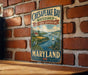A vintage metal sign for Chesapeake Bay Watershed Cultural Landscape, featuring a scenic illustration of a river, farm, and sailboat, mounted on a brick wall.