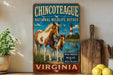 a vintage-style metal sign with a painting of two horses and a lighthouse, placed on a wooden surface next to a potted plant and some lemons.