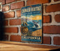 A metal sign with a colorful illustration of a whale, a basket, and a sunset over the ocean, mounted on a brick wall.