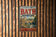 A vintage metal sign that reads "City of Bath United Kingdom" with a picture of a cityscape and a river, mounted on a wooden wall.