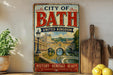 A vintage-style metal sign that reads "CITY OF BATH UNITED KINGDOM HISTORY - HERITAGE - BEAUTY" is displayed on a shelf next to a potted plant and a wooden cutting board.
