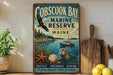 A vintage-style metal sign for Cobscook Bay, Maine, featuring an eagle, a seal, and a boat, along with the text "Cobscook Bay Marine Reserve Maine" and "Protect, Preserve, Inspire".