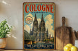 A vintage-style poster of the Cologne Cathedral in Germany, featuring the cathedral's spires and a bridge, is displayed on a wall next to a wooden cutting board and some potted plants.