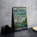 A vintage-style sign for the Connecticut National Estuarine Research Reserve, featuring a seagull and a lighthouse, is displayed on a gray countertop next to some black camera lenses.