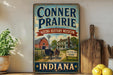 a vintage-style sign for the Conner Prairie Living History Museum in Indiana, with a farm scene and a horse-drawn carriage depicted on the sign.