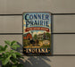 A vintage-style metal sign for the Conner Prairie Living History Museum in Indiana, featuring an illustration of a horse-drawn carriage and a red barn.