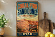 A vintage-style sign for Coral Pink Sand Dunes State Park in Utah, featuring a desert landscape with mountains and a wooden sign that reads "Nature's Pink Paradise Awaits".