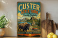 A vintage-style metal sign for Custer State Park in South Dakota, featuring a scenic landscape with a buffalo and a car driving on a road.