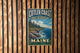 A vintage metal sign for Cutler Coast Public Reserved Land in Maine, featuring a scenic coastal landscape with pine trees, rocky cliffs, and the ocean.