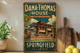 A vintage-style metal sign for Dana Thomas House in Illinois, featuring an illustration of the house and the text "Springfield Illinois" on a wooden shelf.